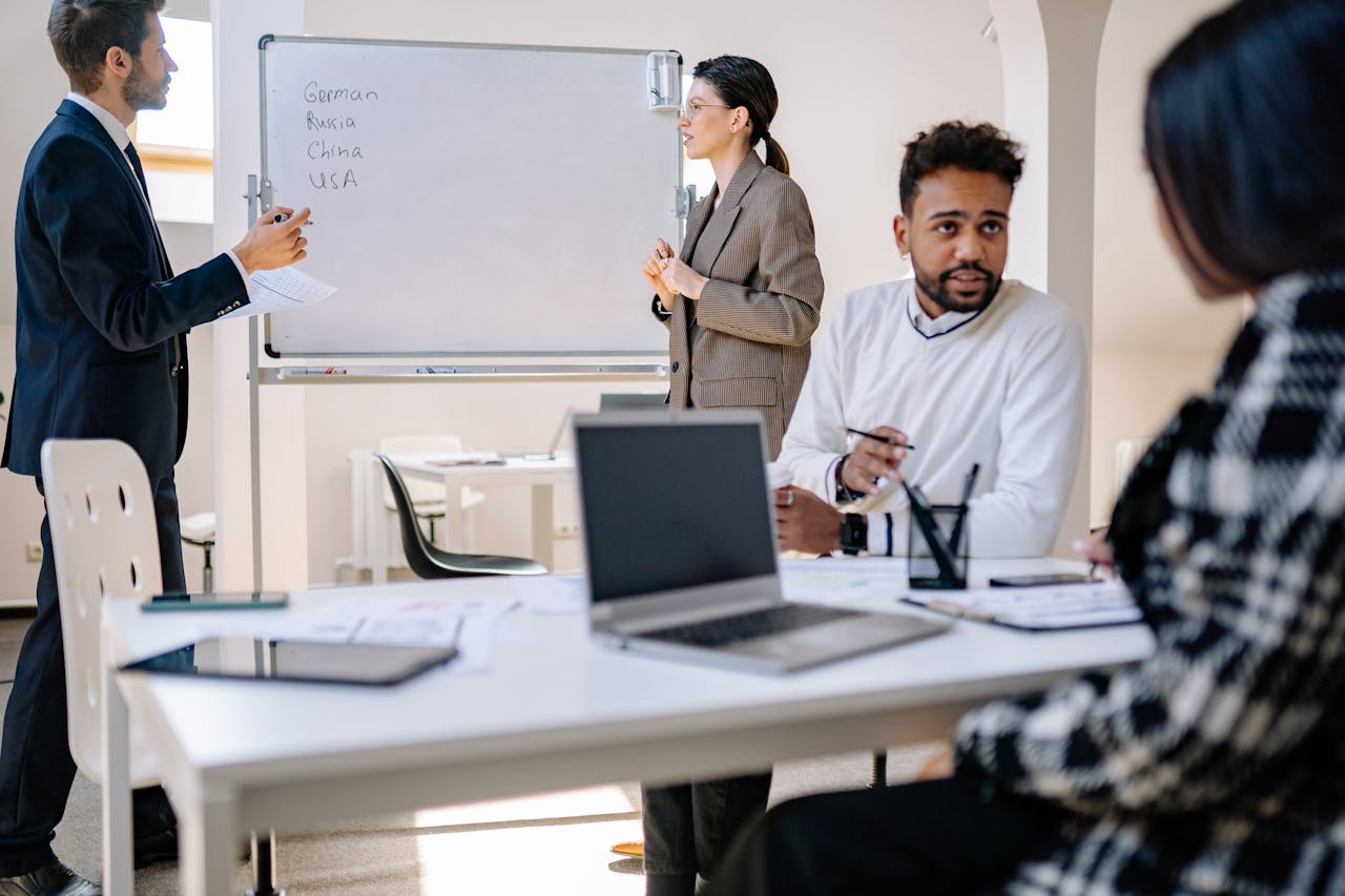 A diverse group of professionals engaged in a collaborative office meeting with laptops and a whiteboard.